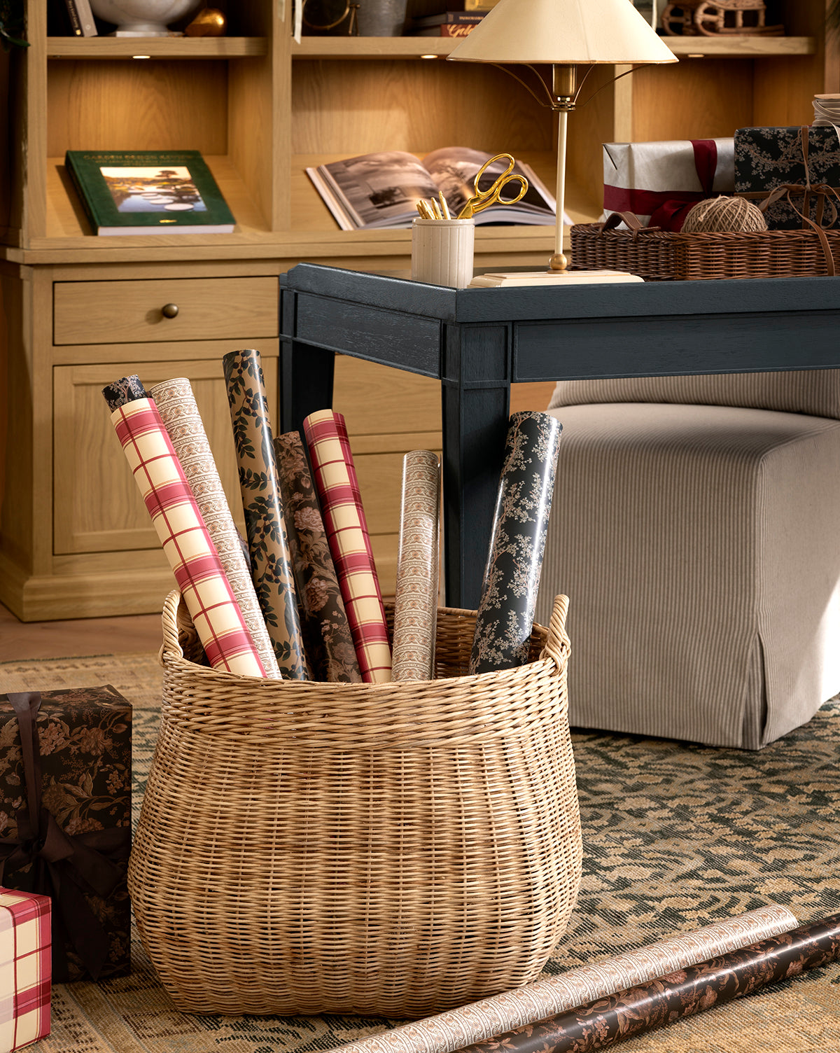 A cozy, well-lit room features a wicker basket filled with Foragers Gift Wrap on a patterned rug near a desk holding wrapped gifts, scissors, and stationary supplies, by McGee & Co.