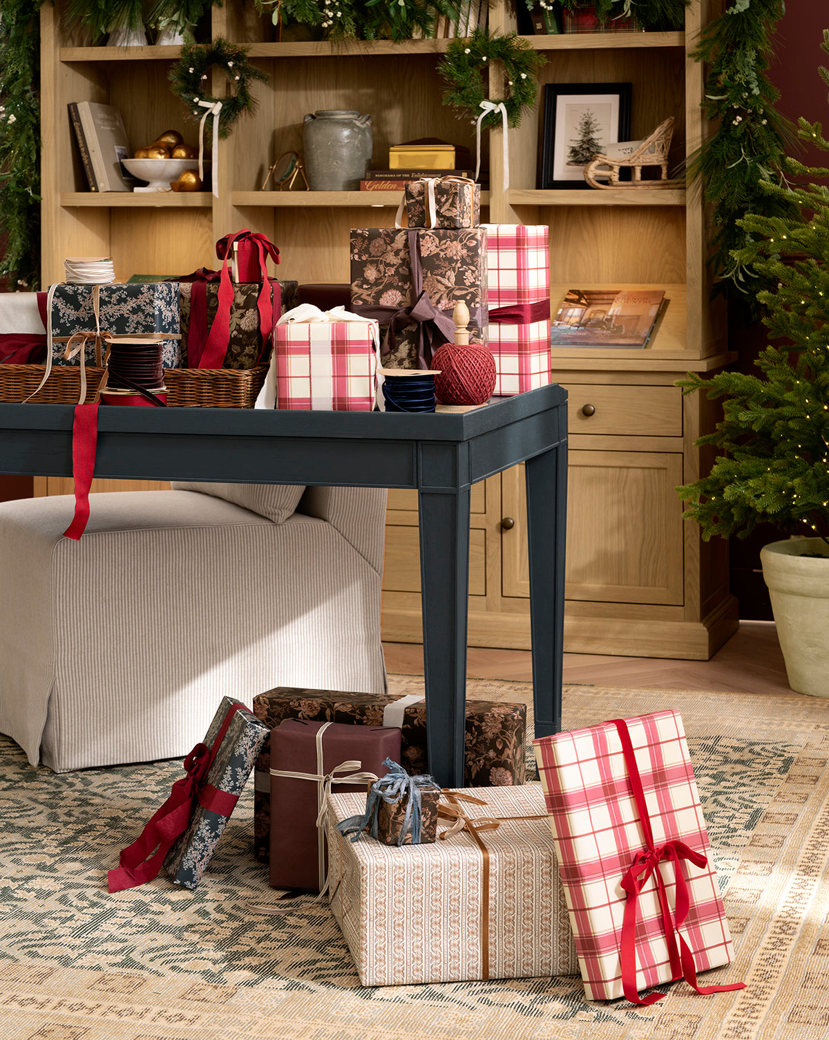 A festive room with a dark-covered table surrounded by gifts wrapped in Highland Plaid Gift Wrap and others, red and pink ribbons, bookshelves adorned with garland, and a Christmas tree in the corner, by McGee & Co.