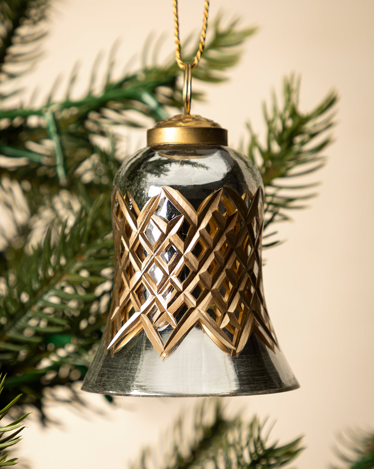 The Hand-Painted Mercury Glass Bell Ornament, in silver and gold with a detailed geometric design, hangs from green pine needles—an ideal touch for festive holiday décor, by McGee & Co.