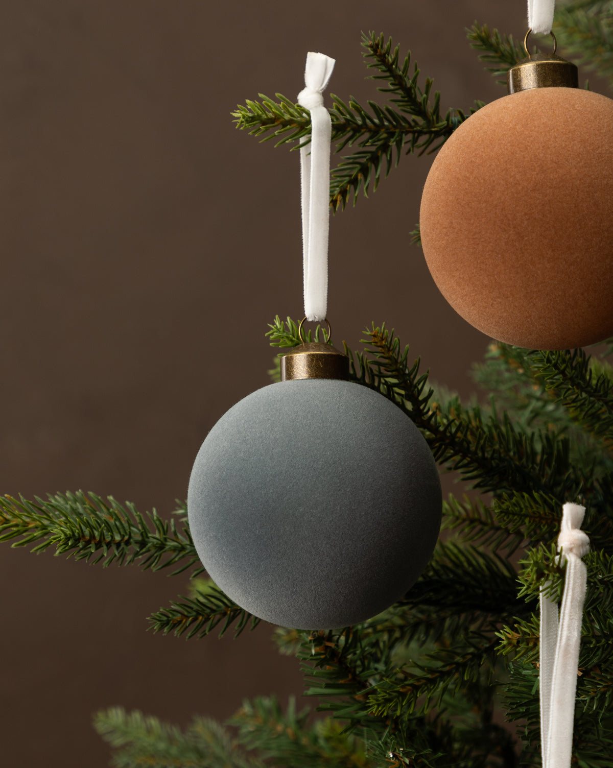 Close-up of a Christmas tree featuring the 3" Assorted Flocked Baubles (Set of 4) in brown, rust, and light gray, each ornament hanging from white ribbons among green pine needles, by McGee & Co.