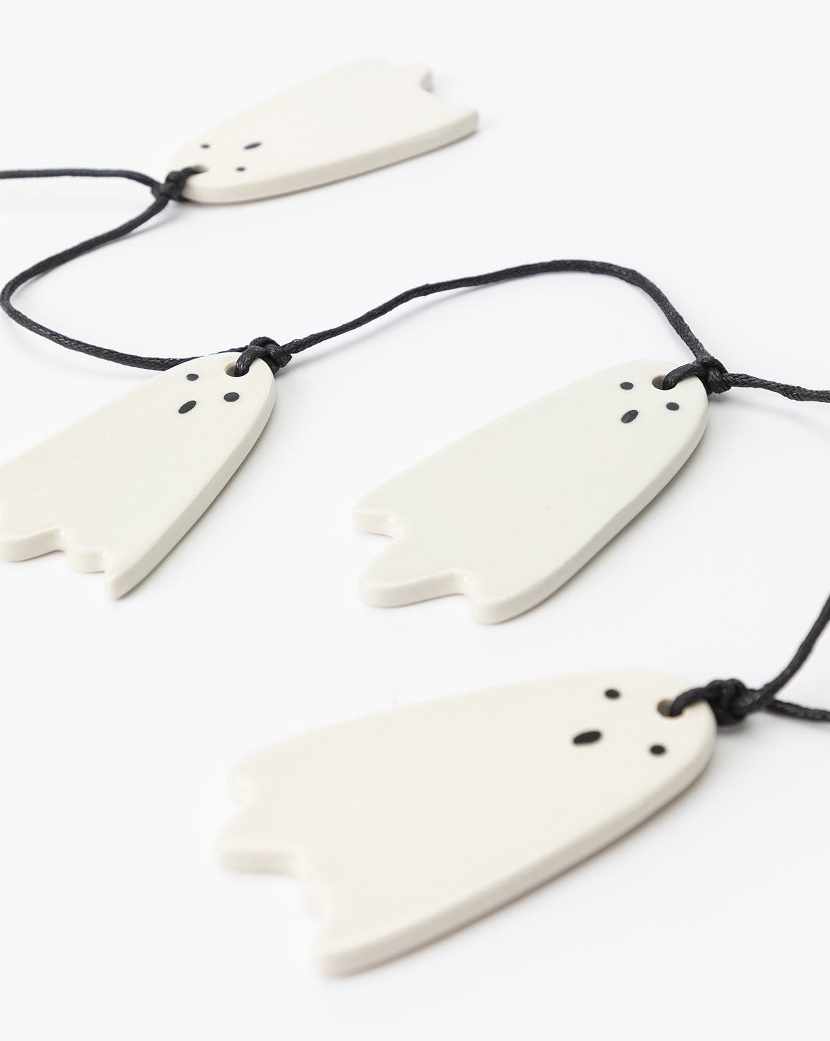 The 3 Ceramic Ghost Garland features four white ceramic ghosts with hand-detailed faces and black eyes and mouths, hanging on black strings—perfect for adding charm to your Halloween decor, by McGee & Co.
