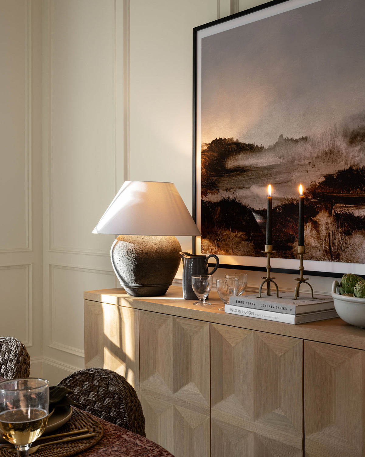 A cozy sideboard with a ceramic lamp, two Claridge Taper Holders, glassware, and books sits below a framed landscape painting, adding elegant mantel decor to the softly lit room.