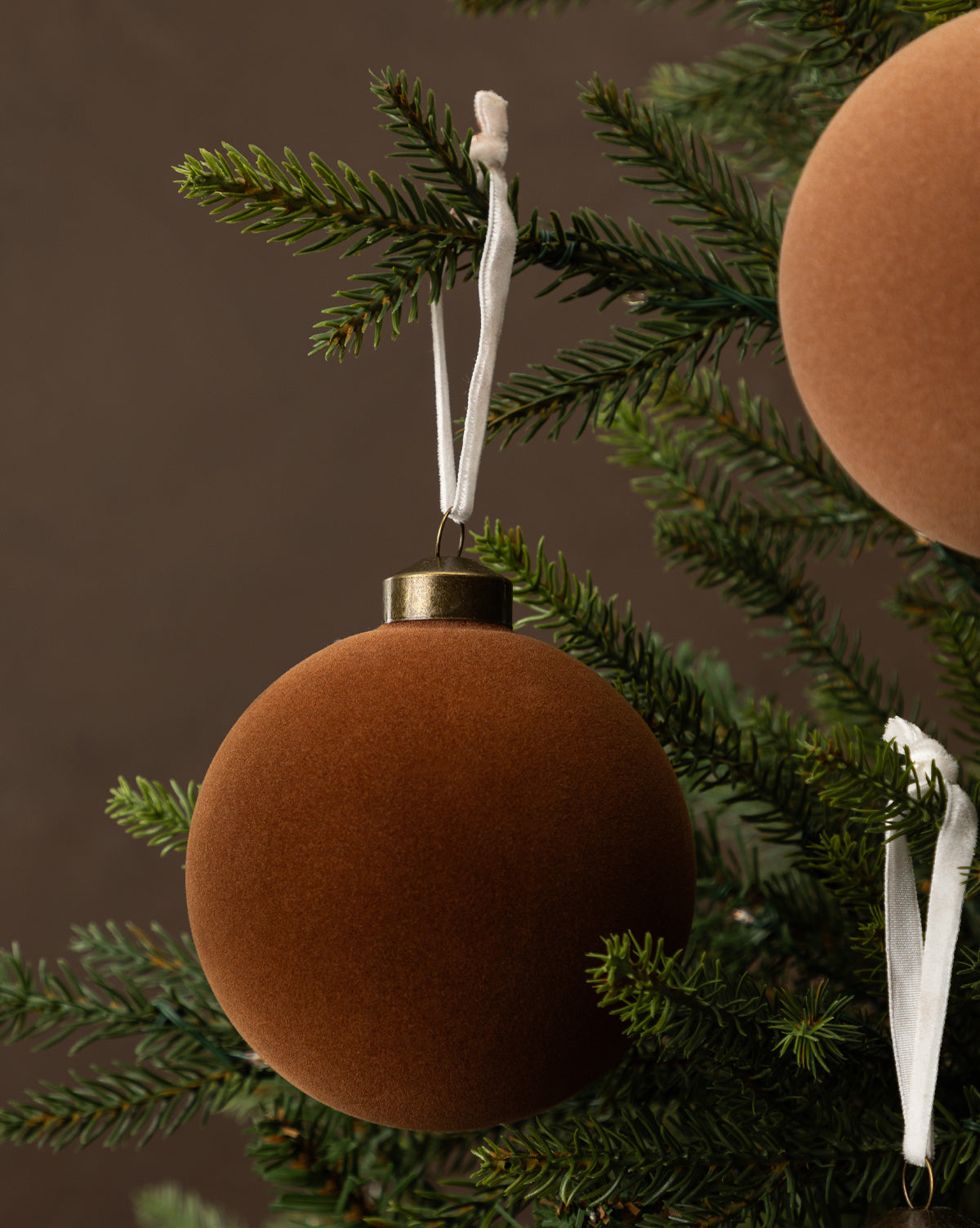 A close-up of a Christmas tree adorned with 4" Assorted Flocked Baubles (Set of 4) in matte blue, brown, and rust, adding festive flair to its green branches, by McGee & Co.