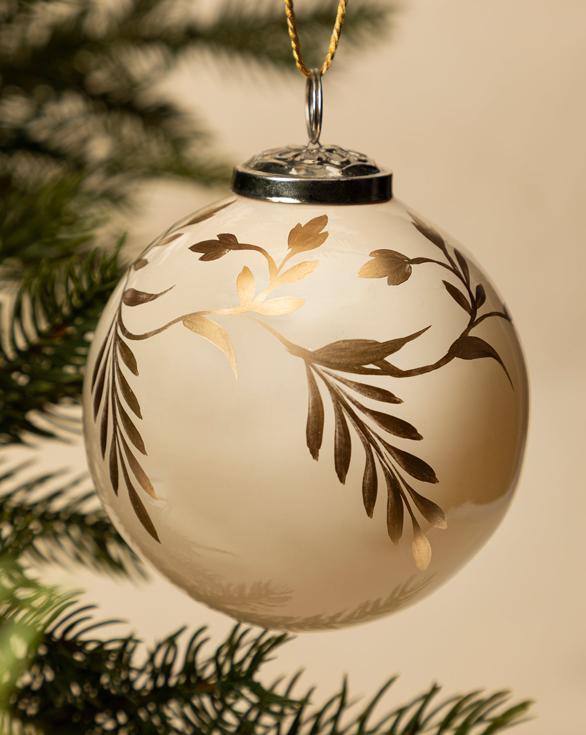 A close-up of the 4" Hand-Painted Vine Glass Bauble, featuring elegant gold leaf patterns, hangs from a pine branch—offering festive inspiration for holiday decorating with unique beaded ornaments, by McGee & Co.