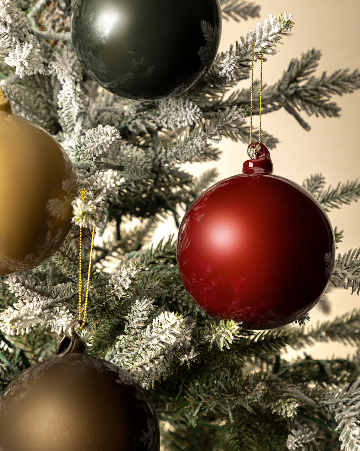 Close-up of a Christmas tree with snow-dusted branches, decorated for the holidays with 4" High Gloss Glass Baubles (Set of 4) in red, green, and gold, hanging from shimmering gold strings, by McGee & Co.