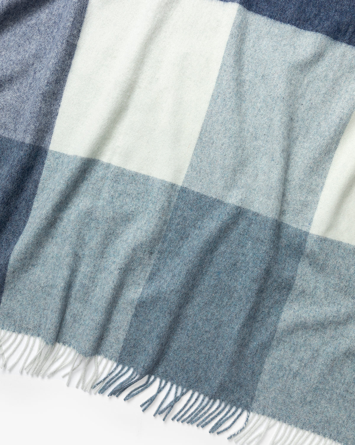 The Abner Throw by Fibre is a soft blue plaid blanket with a large blue, gray, and white pattern, finished with fringed edges and crafted from recycled alpaca wool for an eco-friendly touch. McGee & Co.