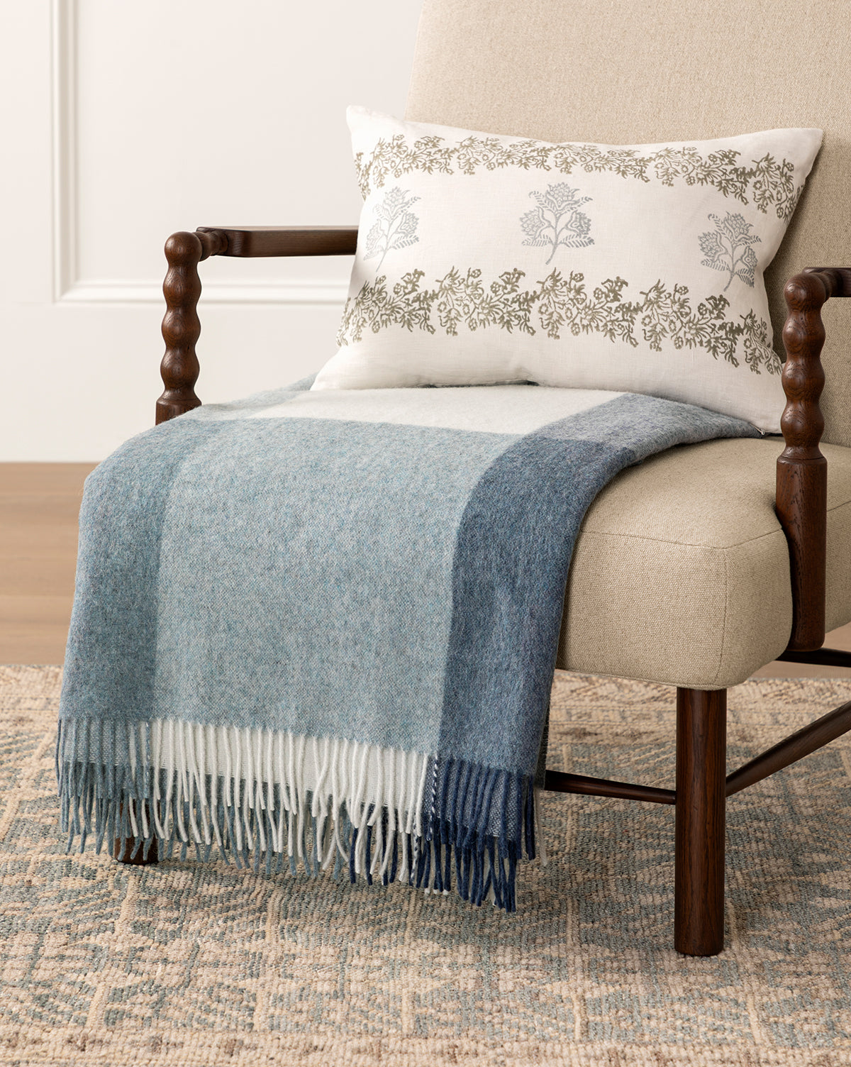 A beige armchair with turned wooden arms is draped with the eco-friendly Abner Throw in blue and gray stripes, and topped with a white pillow featuring floral embroidery, all set on a textured rug, by McGee & Co.