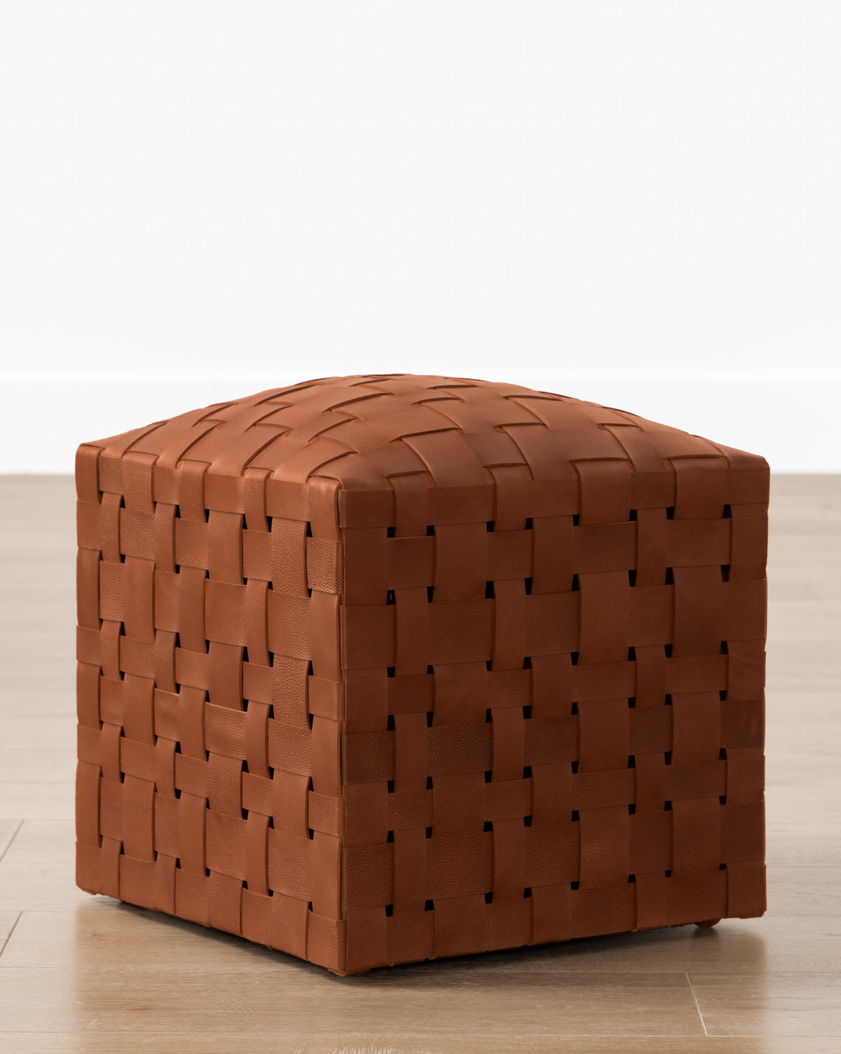 The Ackley Leather Ottoman, featuring a handmade brown leather weave, rests on a light wood floor against a plain white wall, adding stylish extra seating.