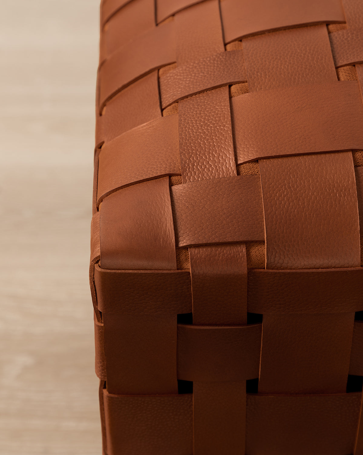 Close-up of an Ackley Leather Ottoman corner, highlighting brown, wide leather strips woven in a natural textured pattern. Set against a light wood surface background.