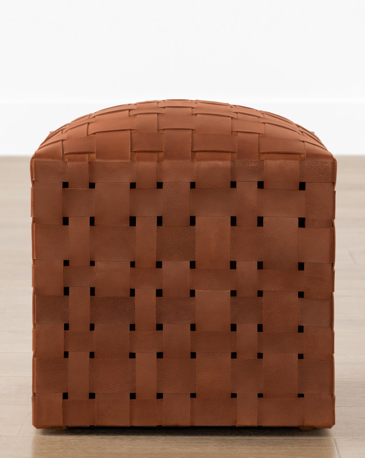 The Ackley Leather Ottoman, handmade in a warm brown weave, adds extra seating on a light wooden floor against a plain white wall.