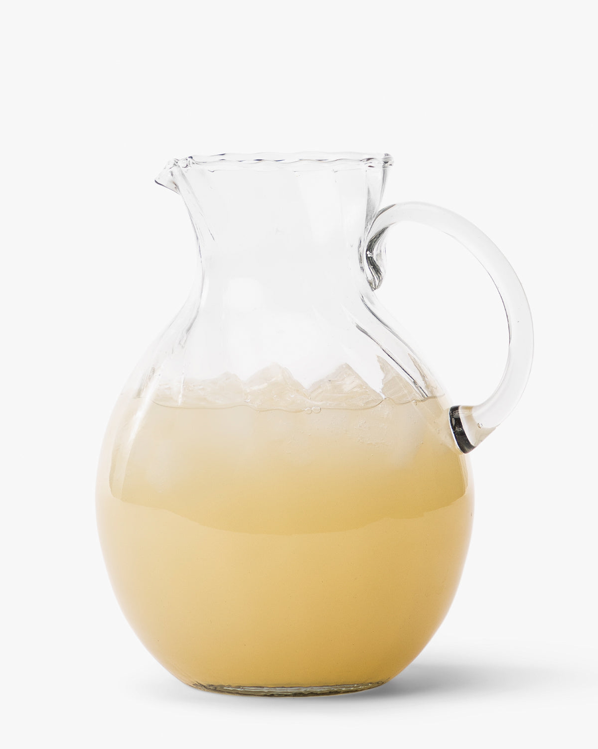 The Adderley Pitcher, a mouth-blown glass piece with a scalloped rim, holds light yellow juice and ice cubes, displayed against a plain white background by McGee & Co.