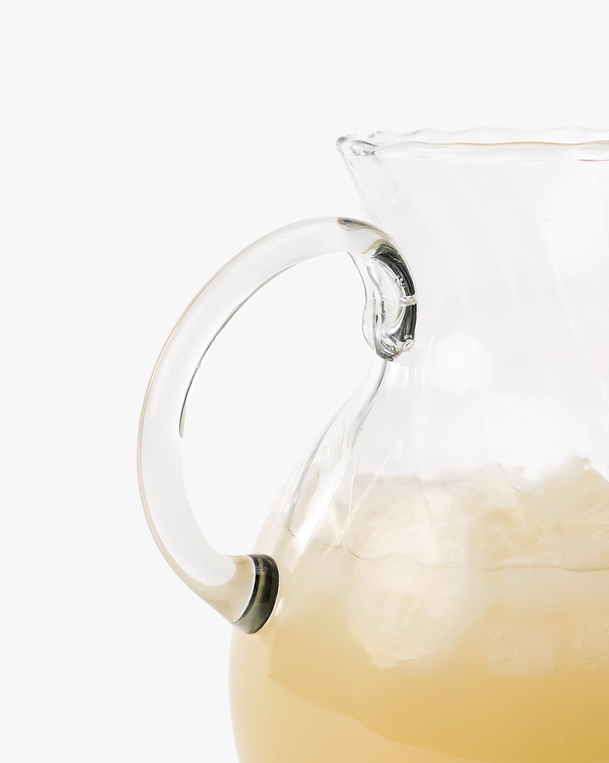 A close-up of the Adderley Pitcher, a mouth-blown glass piece with a scalloped rim, filled with an iced light-colored drink. The focus is on the pitcher’s handle and ice, set against a plain white background, by McGee & Co.