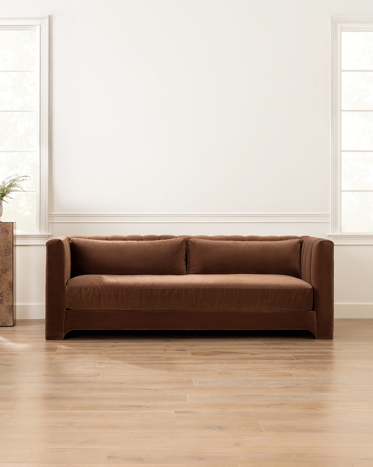 A modern living room showcases the Agatha Sofa 90 in brown velvet against a white wall, bordered by tall windows, by McGee & Co.