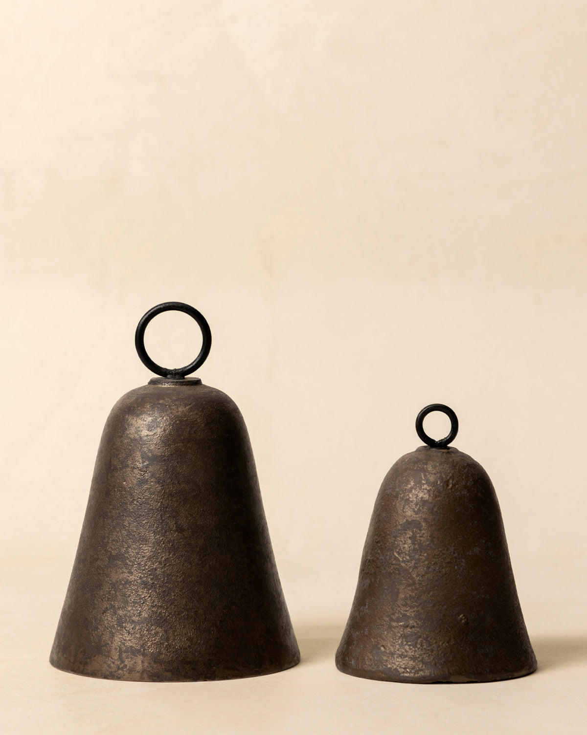 Two Aged Brass Bells with round black handles sit side by side on a light beige surface. The larger and taller bell pairs with a smaller, shorter one, both displaying vintage charm, by McGee & Co.