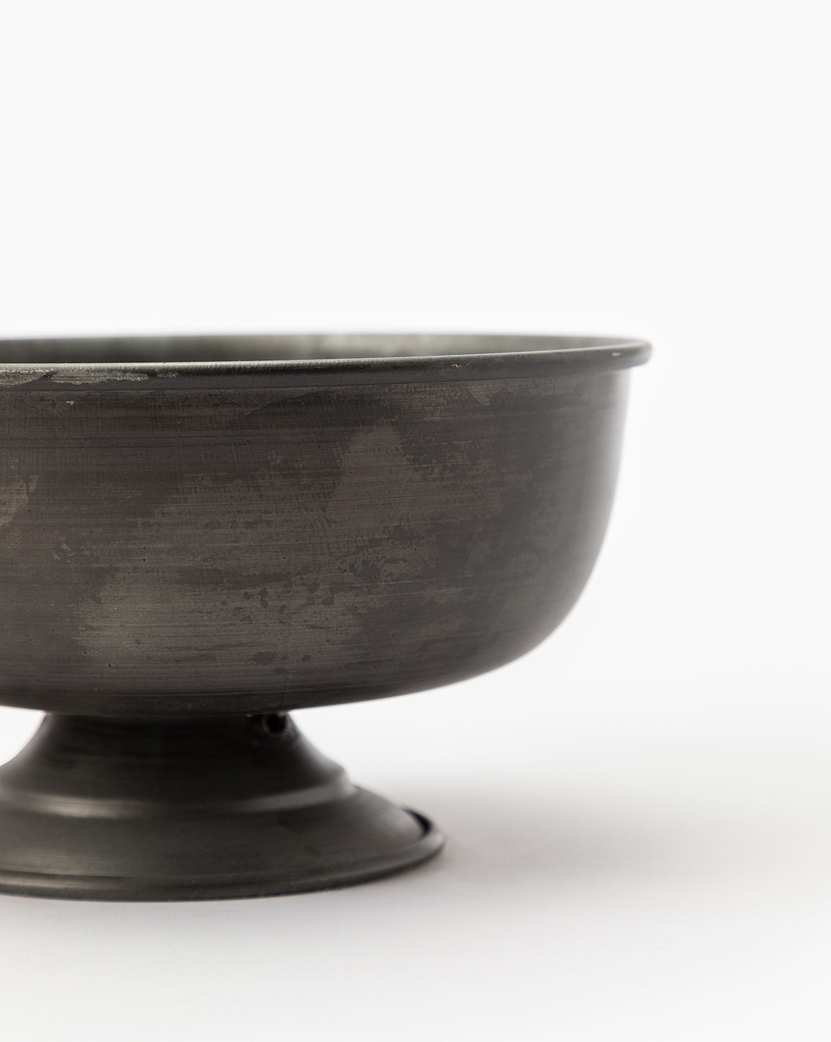 A close-up of the Aged Iron Compote shows its dark gray, matte finish against a plain white background. This rustic vessel is partially visible with its right side out of frame—ideal for floral arrangements.