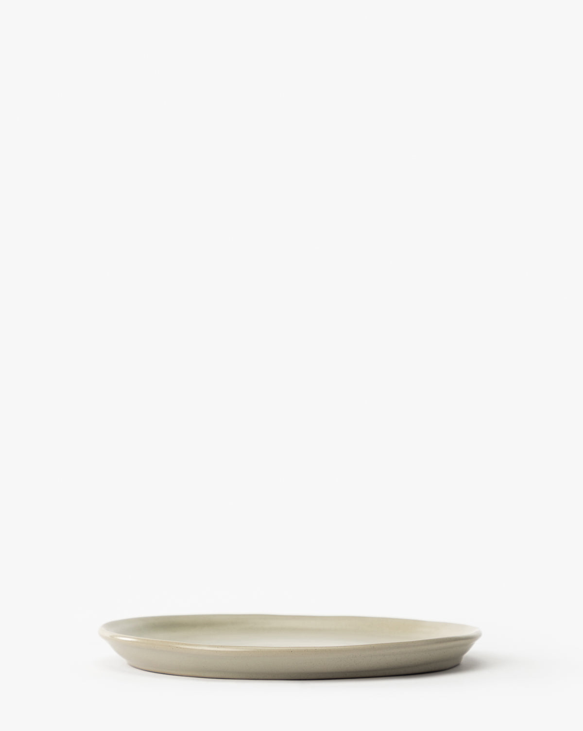 The Aiden Soft Gray Side Plate, a simple round ceramic piece with a light gray glaze, sits on a white background. Viewed from the side, its smooth finish and gently raised edges are highlighted. The plate is empty and elegantly minimalist by McGee & Co.