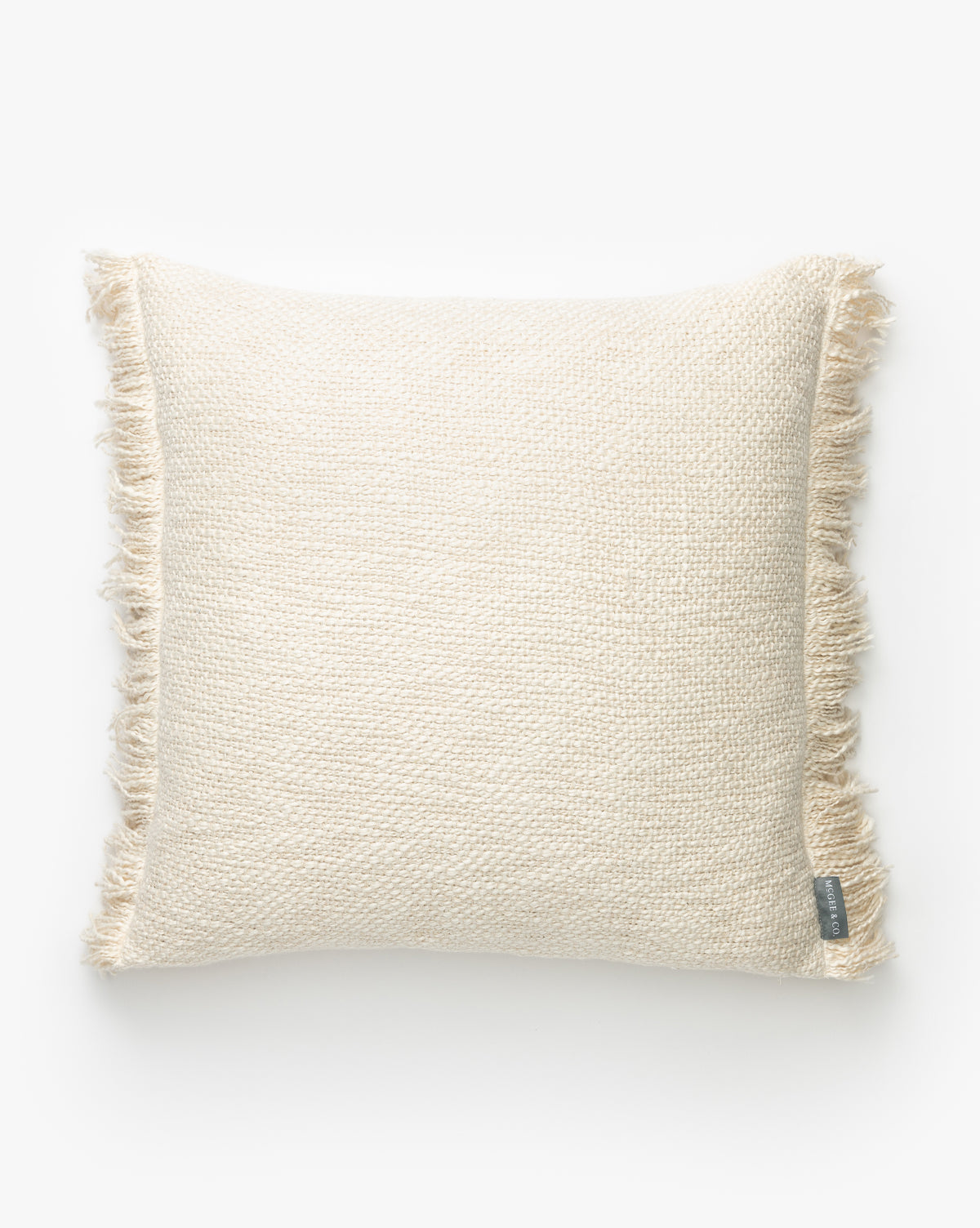 The Aisling Pillow Cover is a square, cream-colored hand-woven pillow cover with a textured design and fringed edges, shown on a plain white background. A small fabric tag appears at the bottom right corner, by McGee & Co.