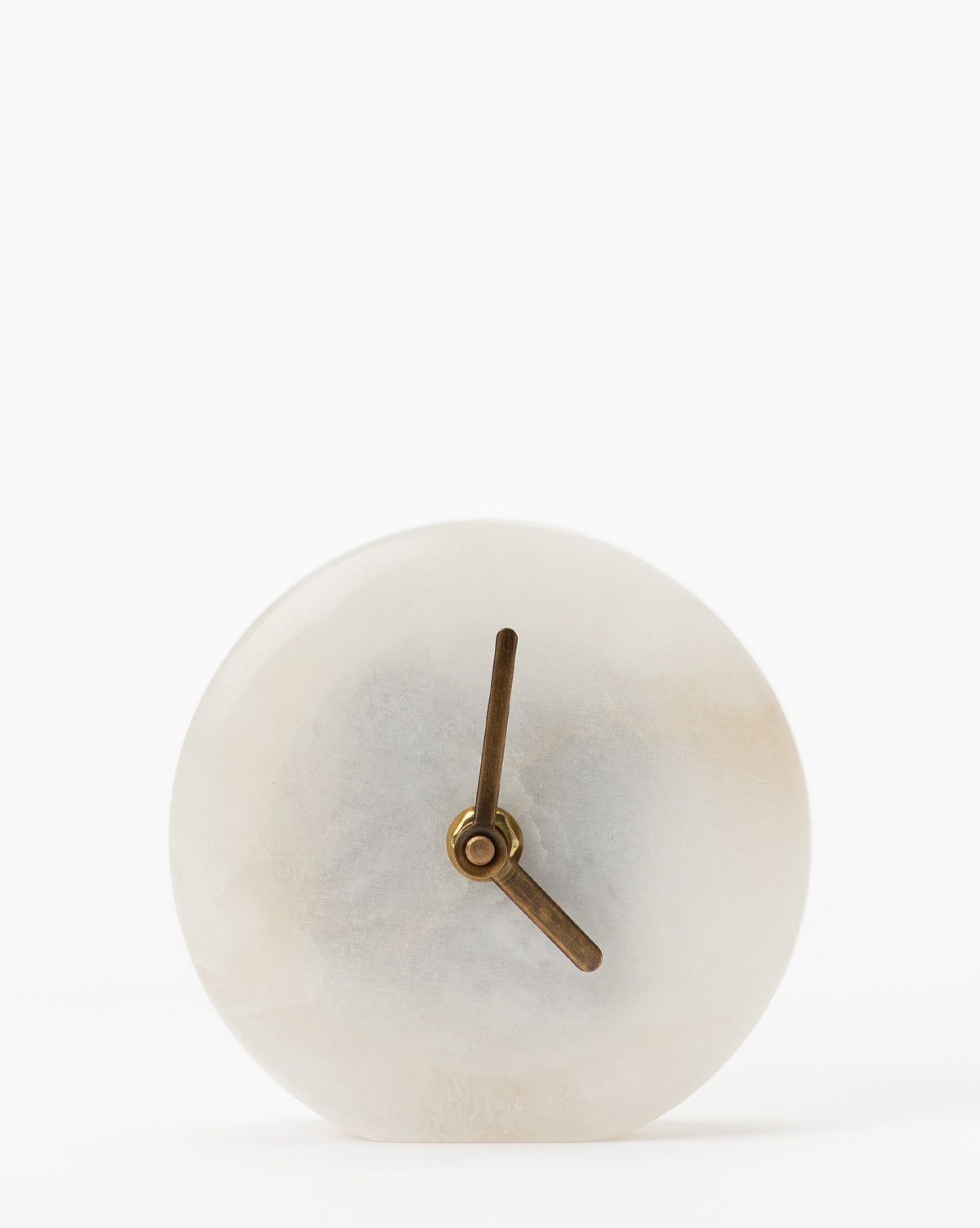 The Alabaster Table Clock features a round, minimalist design with a white marble-like face and simple bronze hands, showcasing sophisticated style against a plain white background.