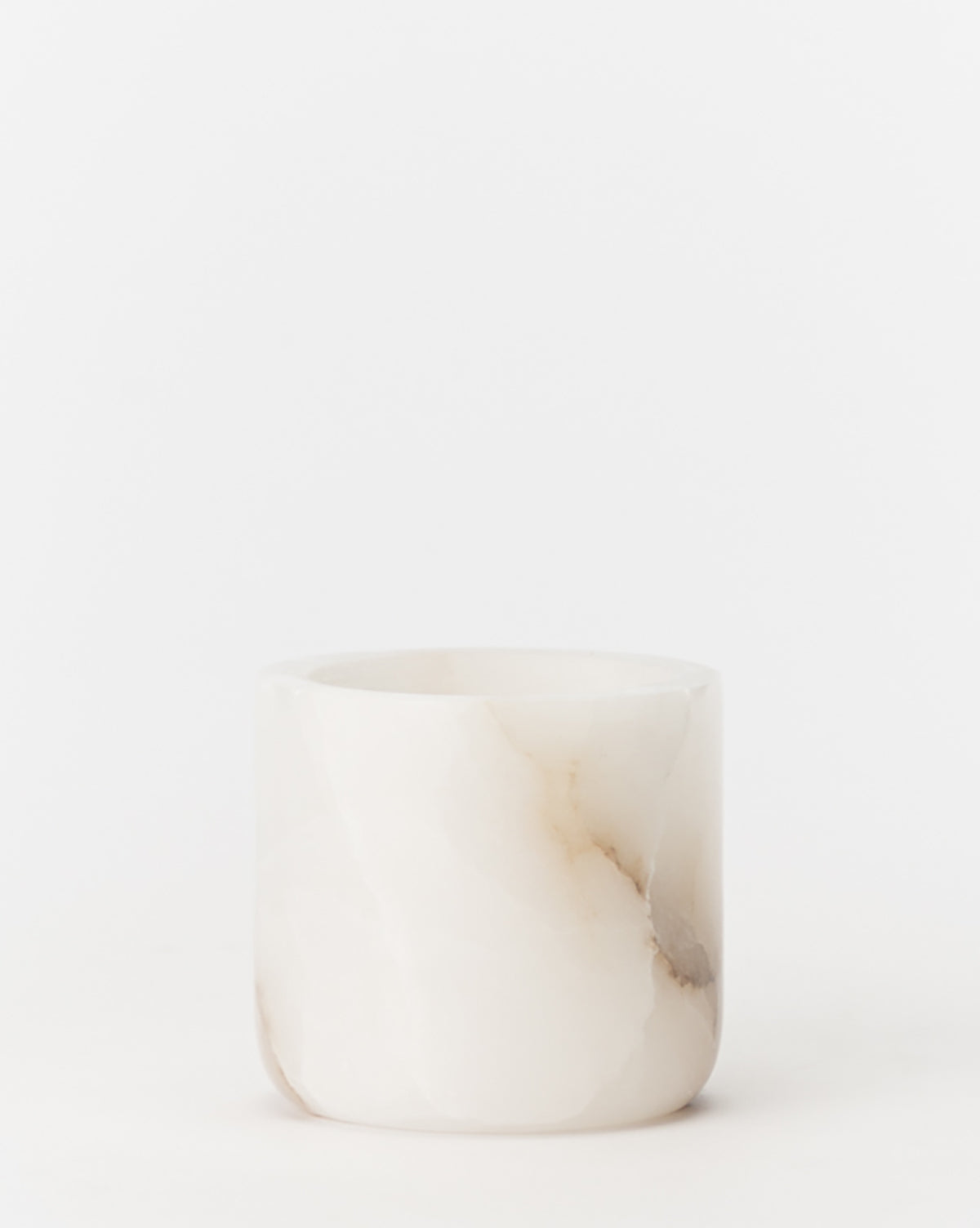 The Alabaster Tea-light Holder is a small, cylindrical white candle holder with delicate brown and beige veining, ideal as a decorative accent, photographed on a plain white background.