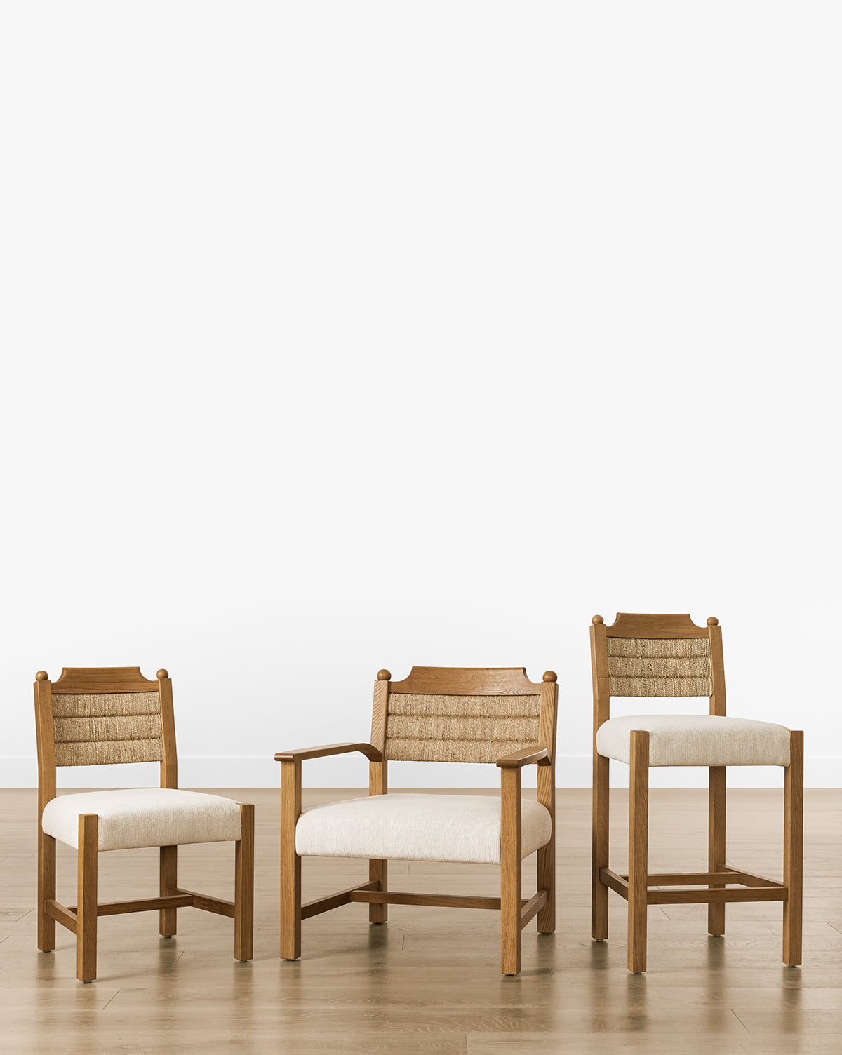 Three wooden chairs, featuring the Makerspalm Alberta Counter Stool in honey oak with seagrass-woven backrests and cream cushions, are set on a light wood floor against a white background. The chairs differ in armrest style and height. McGee & Co.