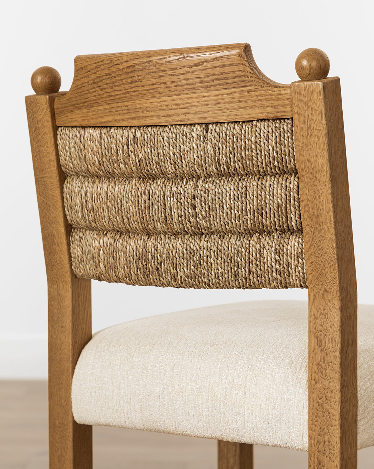 The Makerspalm Alberta Counter Stool features decorative ball finials, a woven seagrass backrest, and a cushioned beige fabric seat. It’s pictured on a light-colored floor against a white wall. McGee & Co.