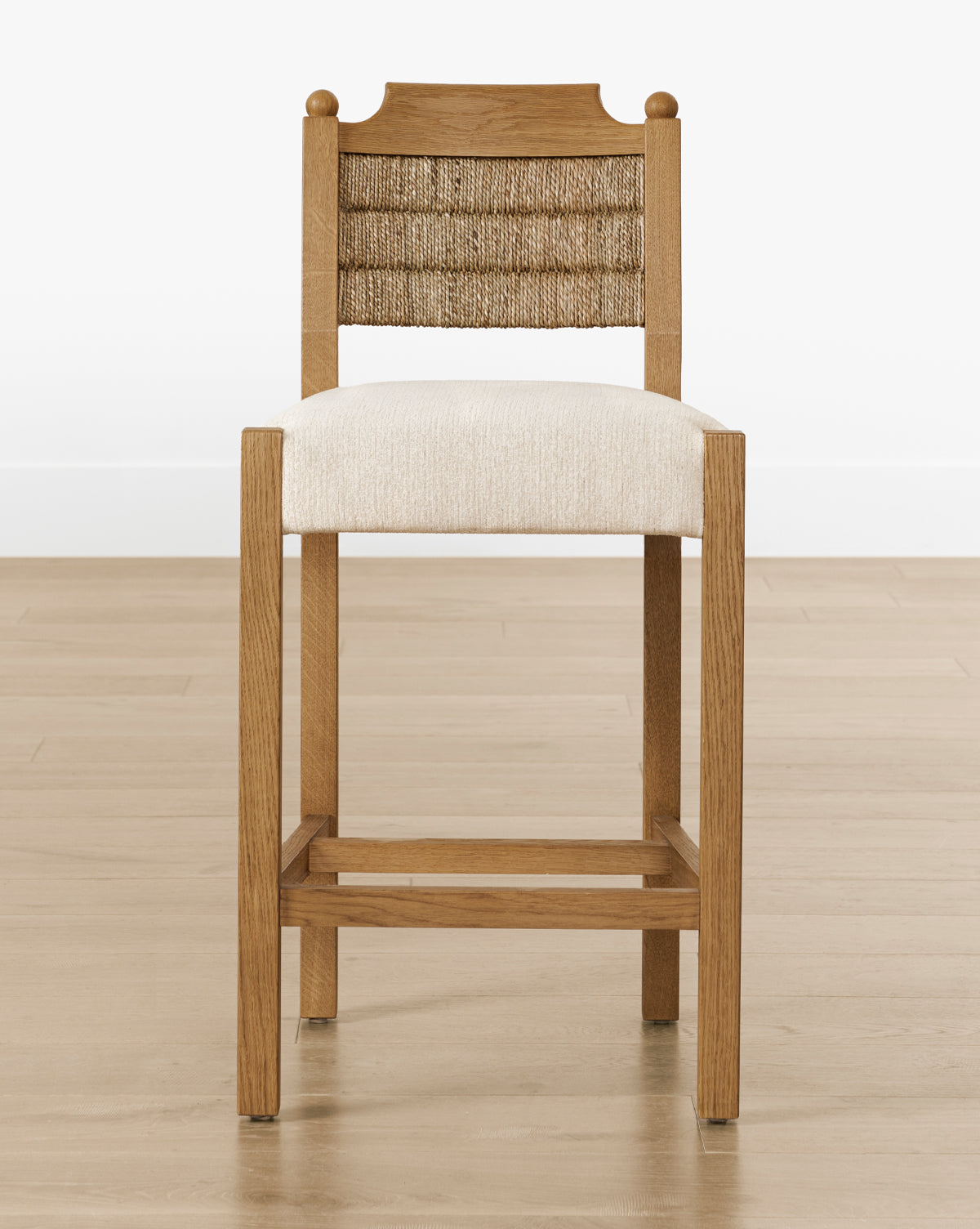 The Makerspalm Alberta Counter Stool has a woven seagrass backrest, cushioned beige seat, and honey oak frame, shown on a light wood floor with a plain white wall backdrop. McGee & Co.
