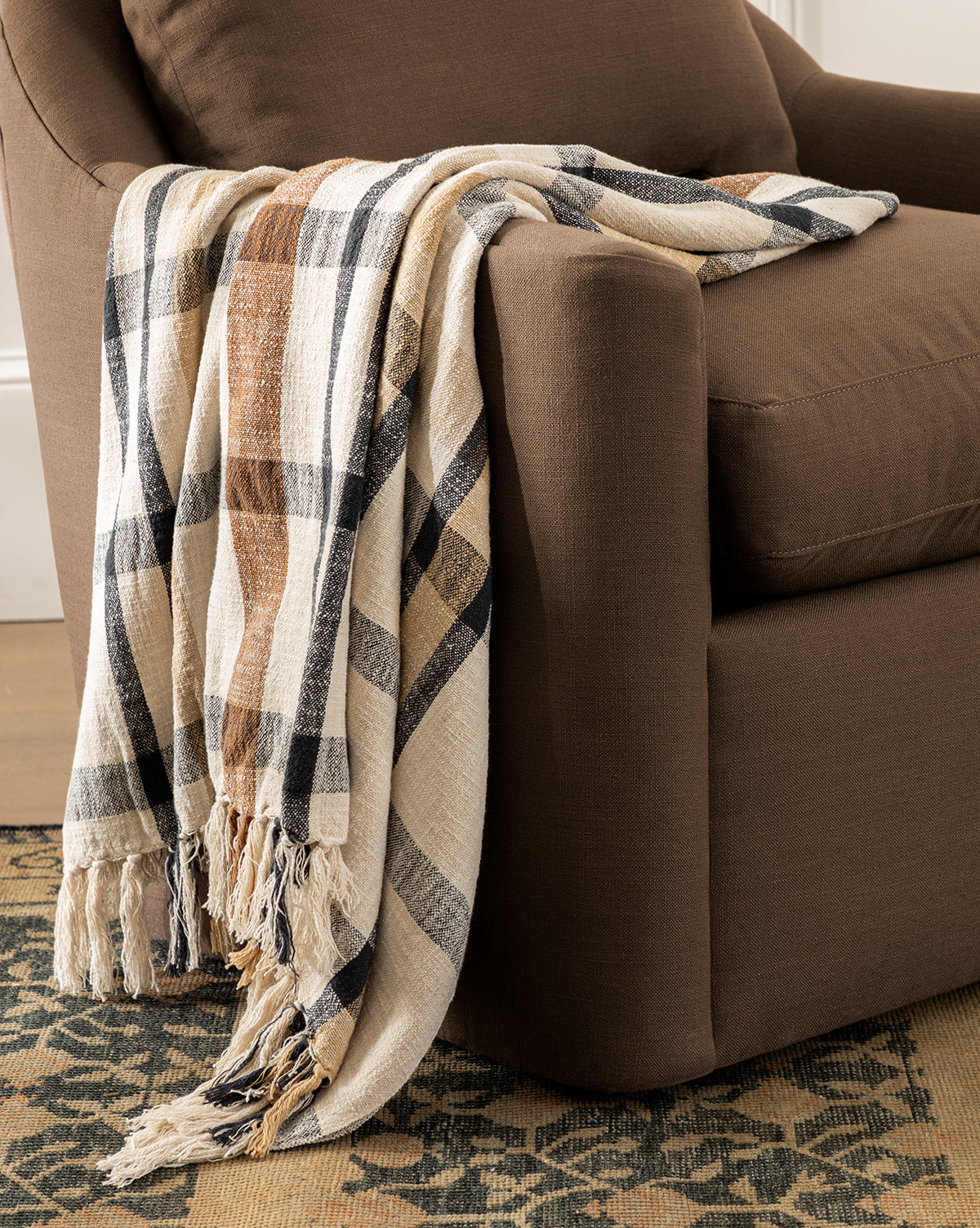 The Raj Albright Plaid Throw, featuring beige, brown, and black stripes, is draped over a brown upholstered armchair. This cozy cotton blanket adds warmth and style to the chair atop a patterned rug in a warmly lit room by McGee & Co.