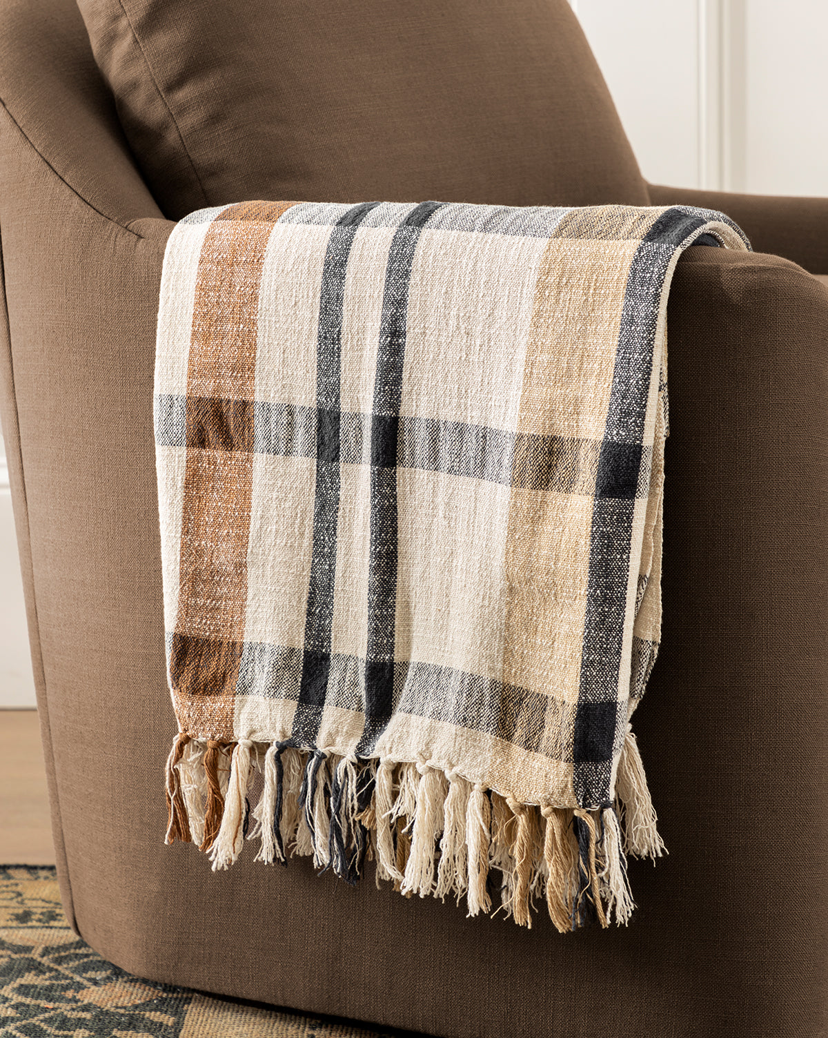 A brown armchair with a Raj Albright Plaid Throw in beige, black, and brown draped over the armrest. The cotton throw features fringed edges and textured fabric for comfort and style, from McGee & Co.