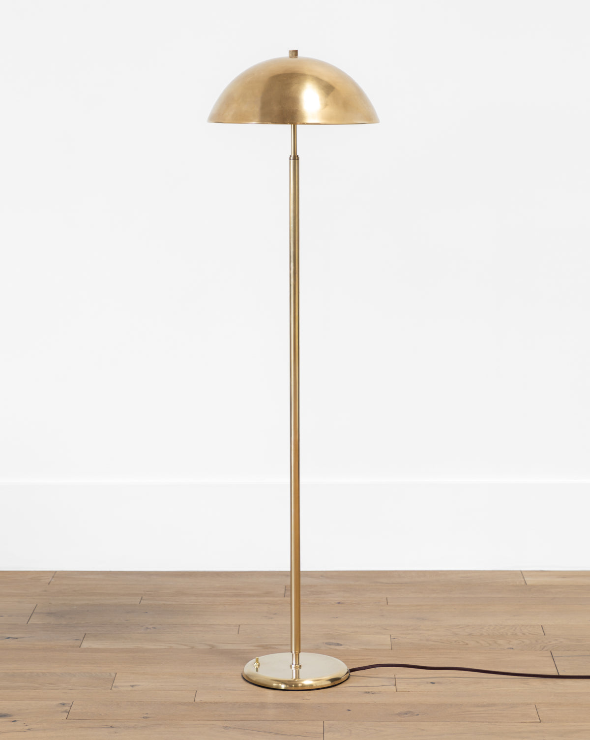 The Makerspalm Aldorno Floor Lamp features a slim pole, wide round shade, and round base on a light wooden floor against a white wall, with a black power cord extending from its base. McGee & Co.