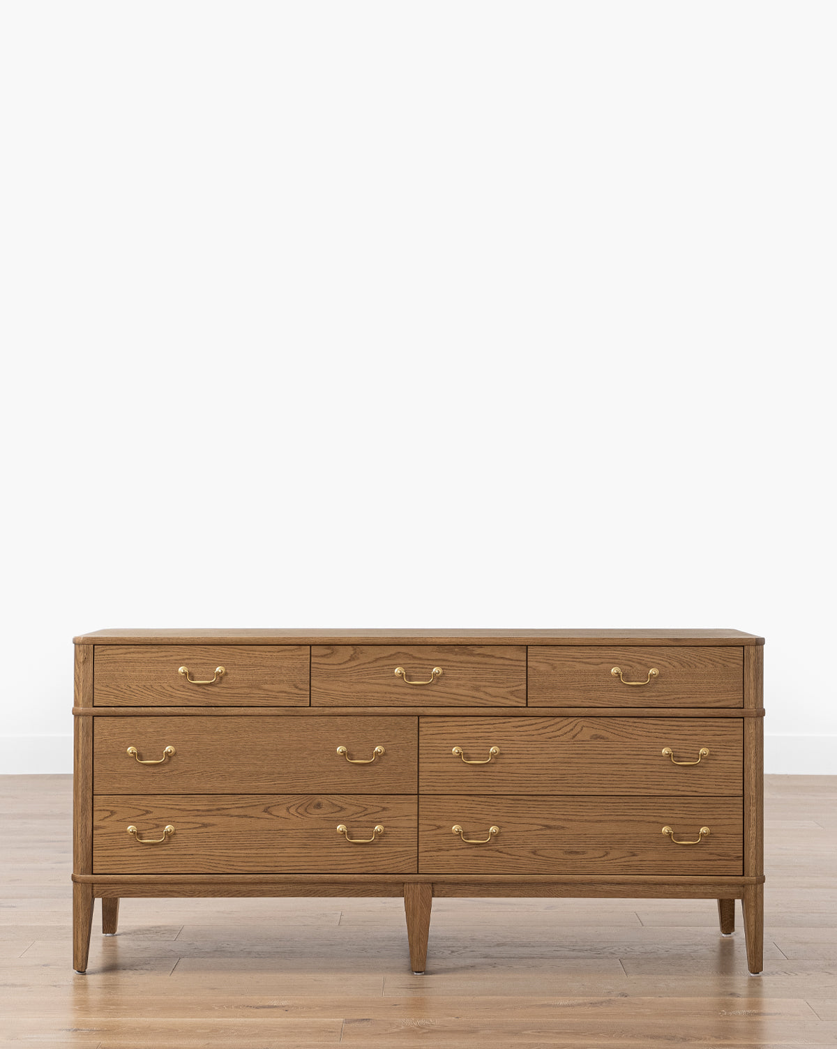The Makerspalm Alessandro Dresser is a honey oak piece with nine soft-close drawers and brass handles, standing on four tapered legs atop a light wood floor against a plain white background, McGee & Co.
