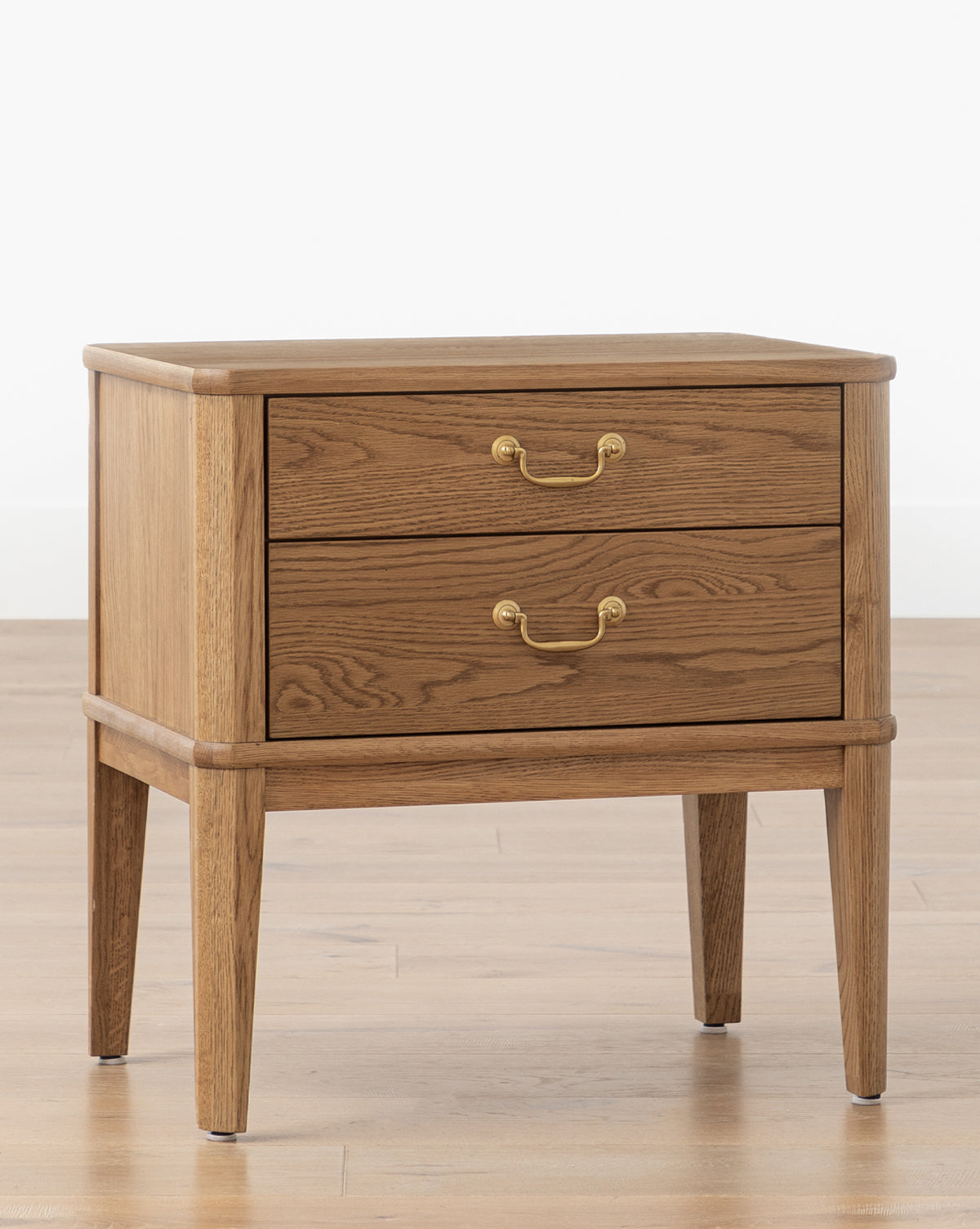 The Alessandra Nightstand, featuring two drawers, brass handles, and tapered legs in elegant honey oak, sits on a light wood floor against a white wall by McGee & Co.