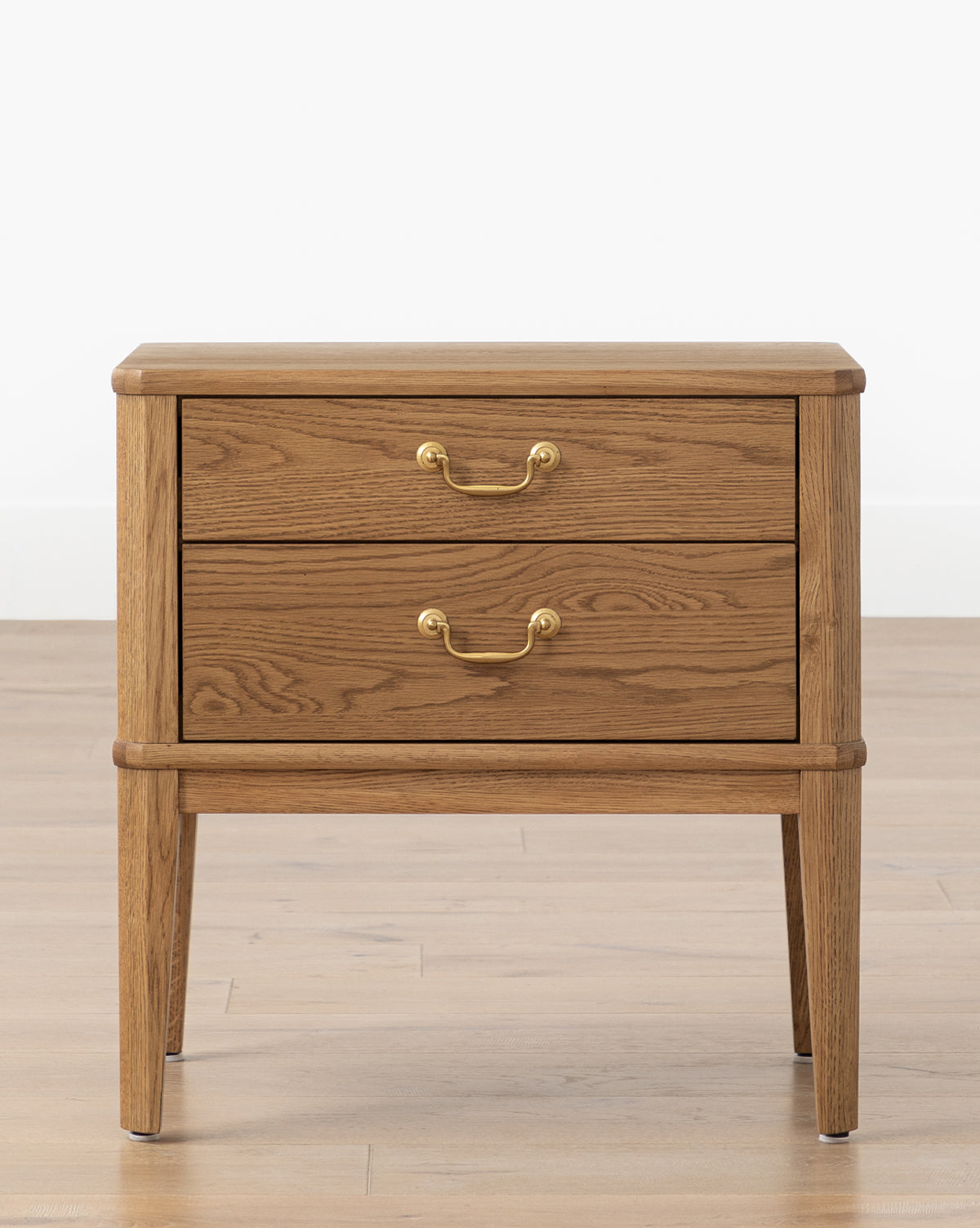 The Alessandra Nightstand by Makerspalm, crafted from solid oak, features two drawers with brass handles and stands on four tapered legs, shown against a light wall and wood floor. McGee & Co.