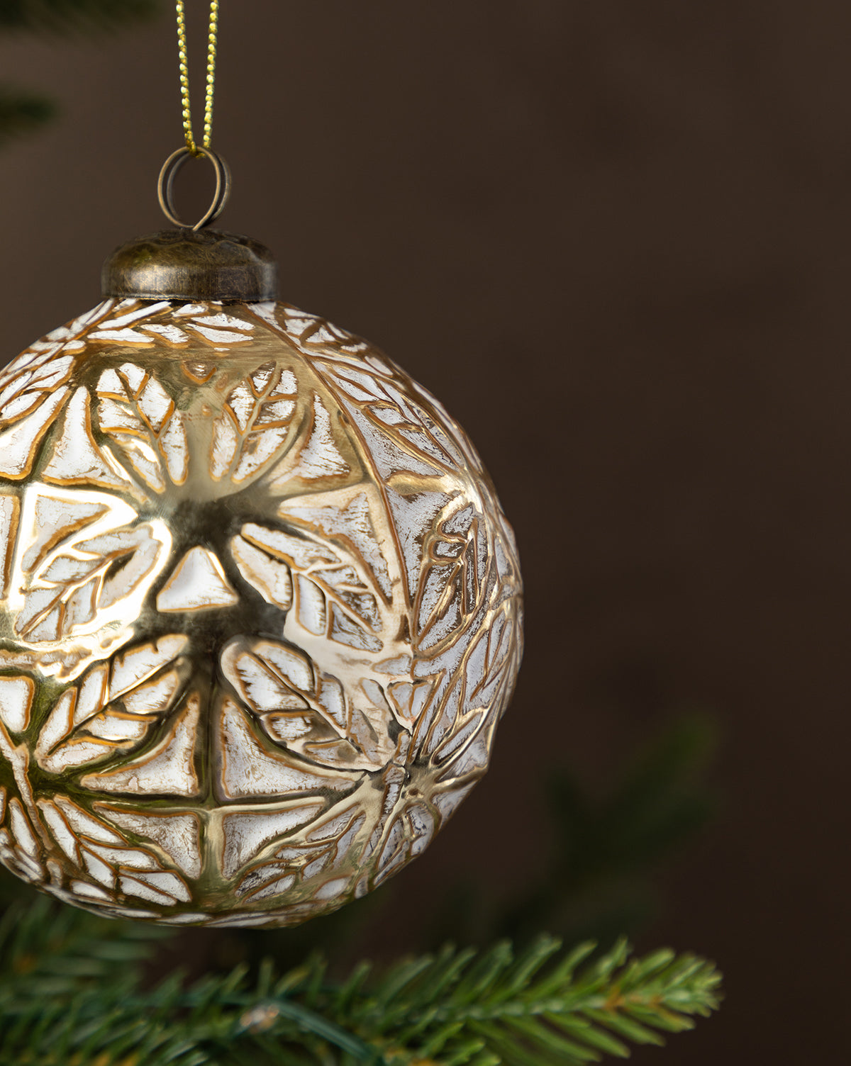 The Alessandro Glass Ornament features a gold leaf and snowflake design, shown in close-up as it hangs from a green Christmas tree branch for festive holiday decor, by McGee & Co.