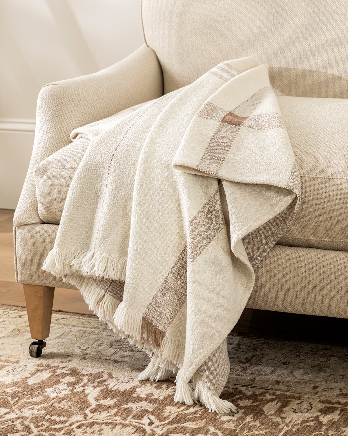 A cream-colored armchair features the ILA HOME FASHIONS Alex Plaid Throw—a beige and tan, fringed blanket—draped over the arm. The chair sits on a patterned rug in a softly lit, cozy room, blending timeless comfort with effortless style from McGee & Co.