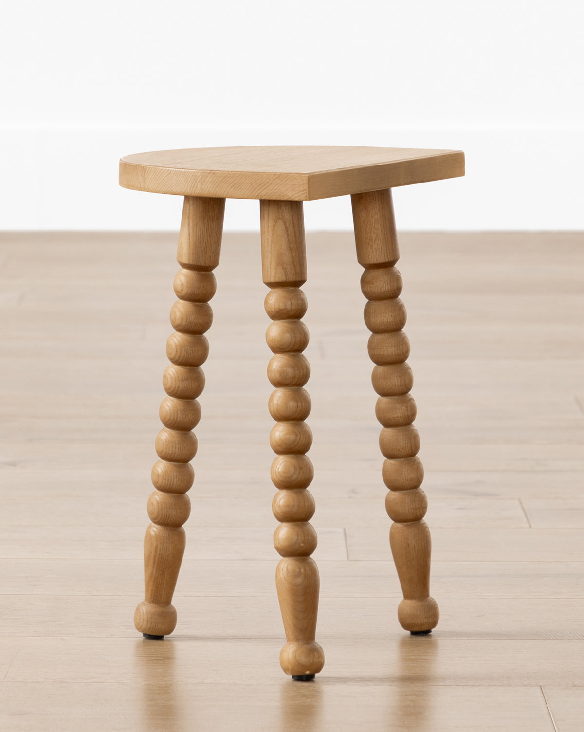 The Alice Honey Wood Stool by Makerspalm features a triangular ash wood seat and spindle legs. Compact and vintage-inspired, it’s perfect as an accent stool for any room. Pictured on a light wood floor against a white background. McGee & Co.
