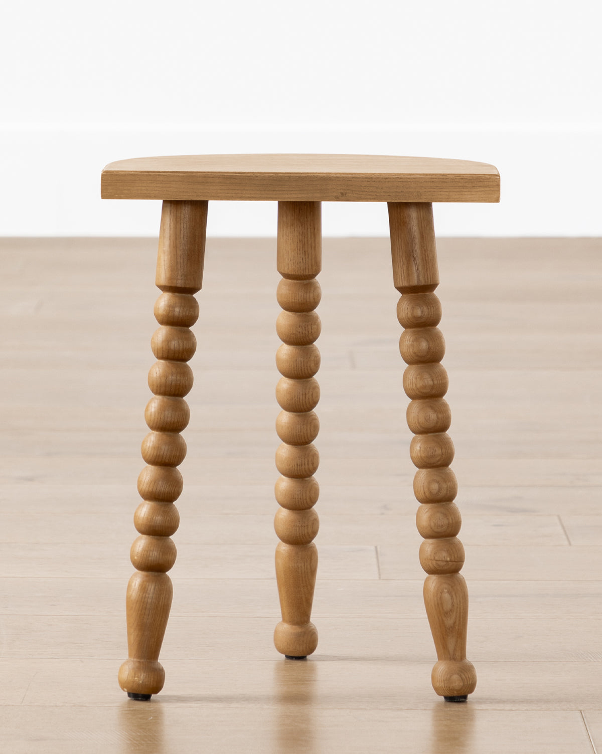 The Alice Honey Wood Stool by Makerspalm features three turned spindle legs and a flat rectangular seat, making it a perfect accent piece. Shown on a light wood floor against a white wall. McGee & Co.