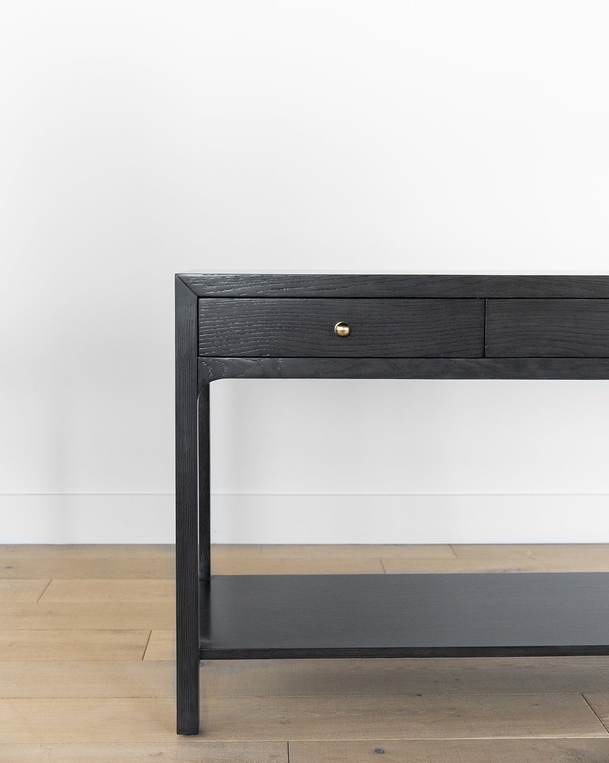 The Allen Black Console is a modern entryway table featuring a black wood finish, a drawer with a brass pull, an open lower shelf, and looks elegant against a white wall on a light wood floor, by McGee & Co.