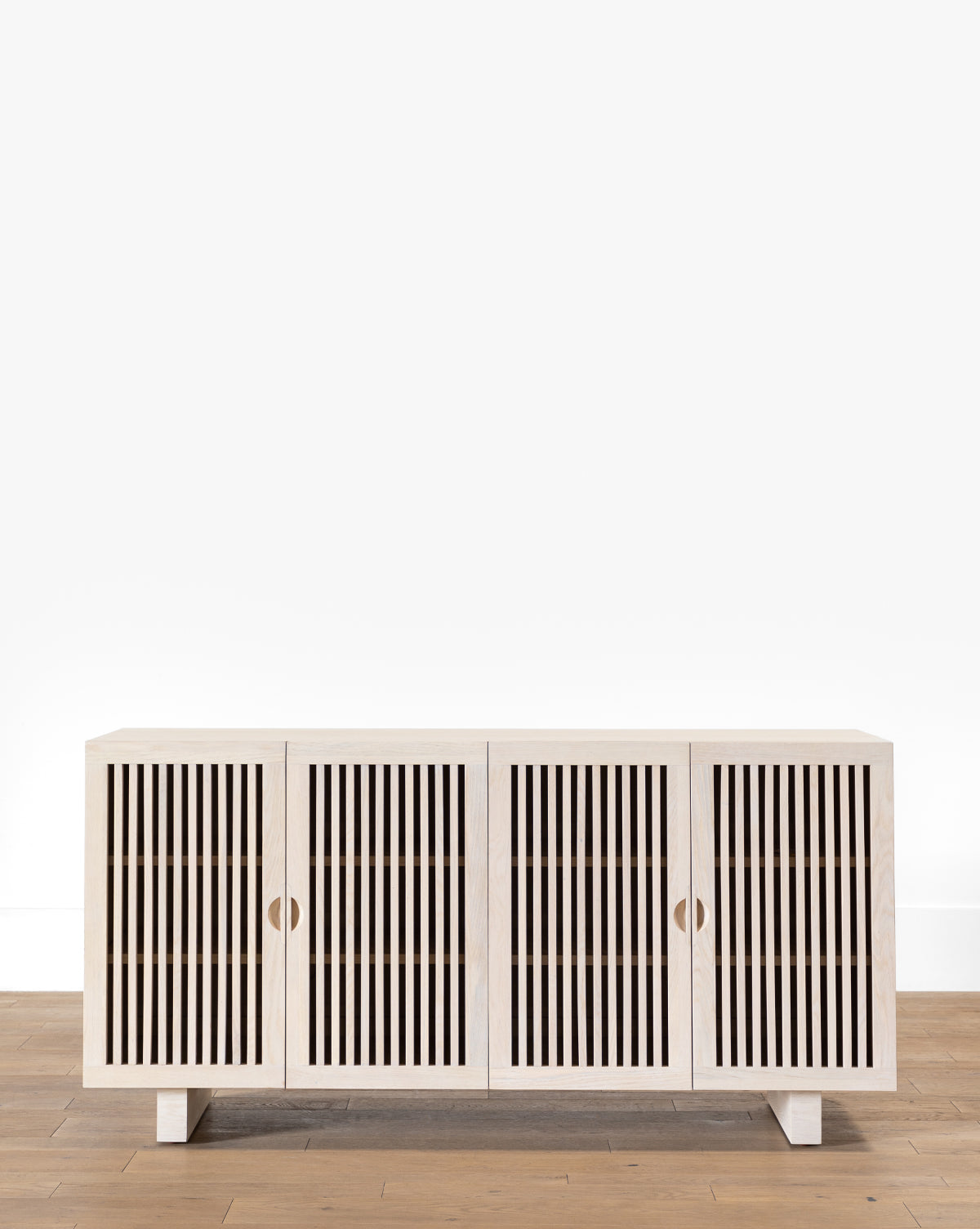 The Makerspalm Alvin Sideboard is a modern solid oak piece with four doors and vertical slats, standing on low square legs, shown here on a light wood floor beside a plain white wall. McGee & Co.