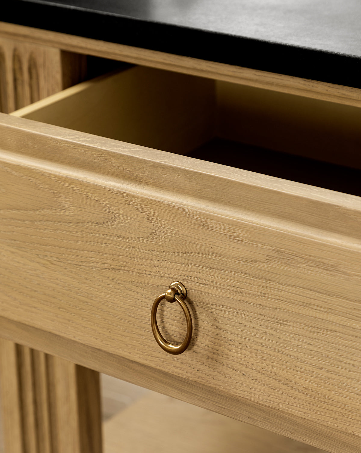A close-up of the Ambrose Nightstand drawer, featuring a brass ring handle and honed granite top. The light-finished wood and empty interior highlight its elegant, vintage-inspired design for the bedroom, by McGee & Co.