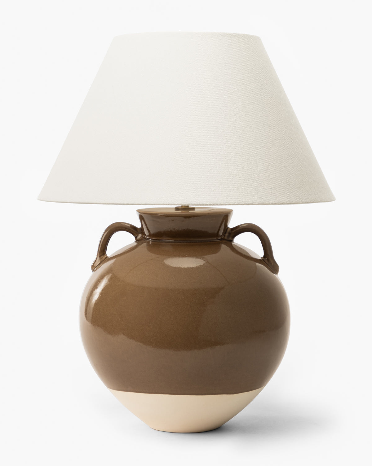 The Makerspalm Amphora Table Lamp features a glossy brown ceramic base with two side handles and a lighter bottom, topped with a wide off-white shade for a classic look. McGee & Co.