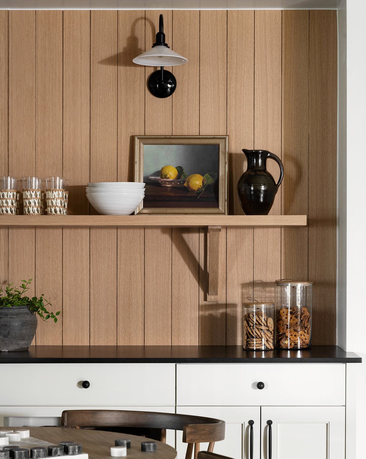A wooden kitchen wall displays a floating shelf with the Bowl of Lemons painting, a white bowl, dark pitcher, glassware, and cookie jars. Below are white cabinets, a plant, and a black countertop for charming kitchen decor.
