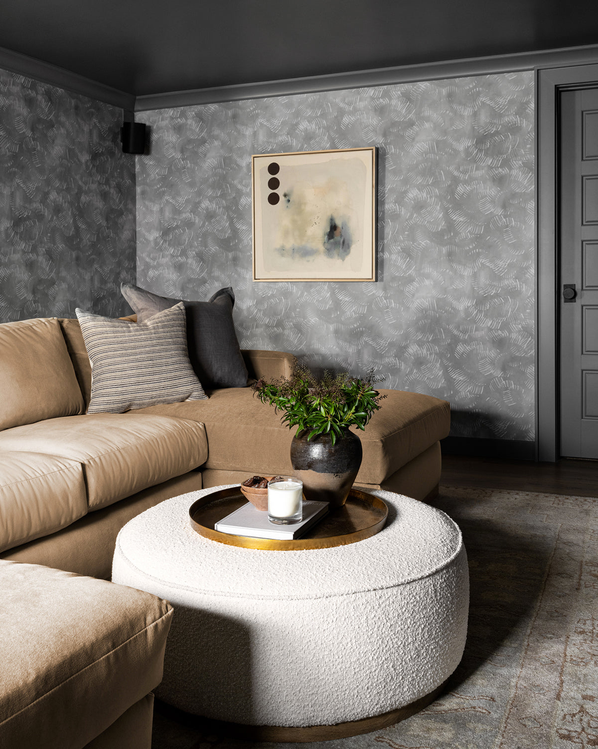 A modern living room features a beige sectional, fabric cushions, gray textured walls, abstract art, and the Dawson Ottoman with a tray for a candle and greenery. A gray door appears in the background, by McGee & Co.