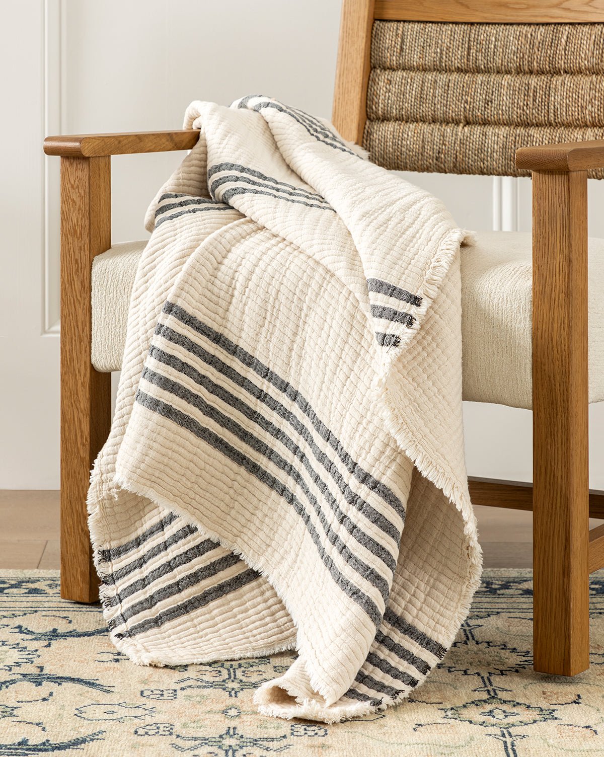 The Creative Co-op Anneliese Striped Throw, featuring a cream and dark gray stripe design, is draped over a cushioned wooden chair on a patterned rug, bringing cozy charm and luxury home decor style to the light-filled room—McGee & Co.