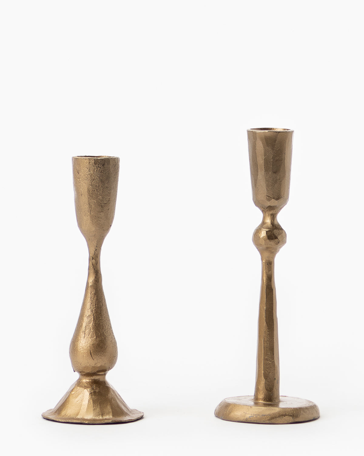 Two mismatched Antique Brass Taper Candle Holders, each hand-formed with a textured surface, stand side by side on a plain white background. Each holder displays a distinct irregular shape and uniquely designed base.