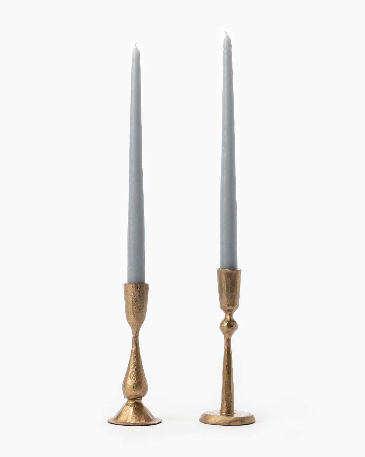 Two tall, slender gray candles are set in mismatched Antique Brass Taper Candle Holders, each with a unique textured design, against a plain white background.