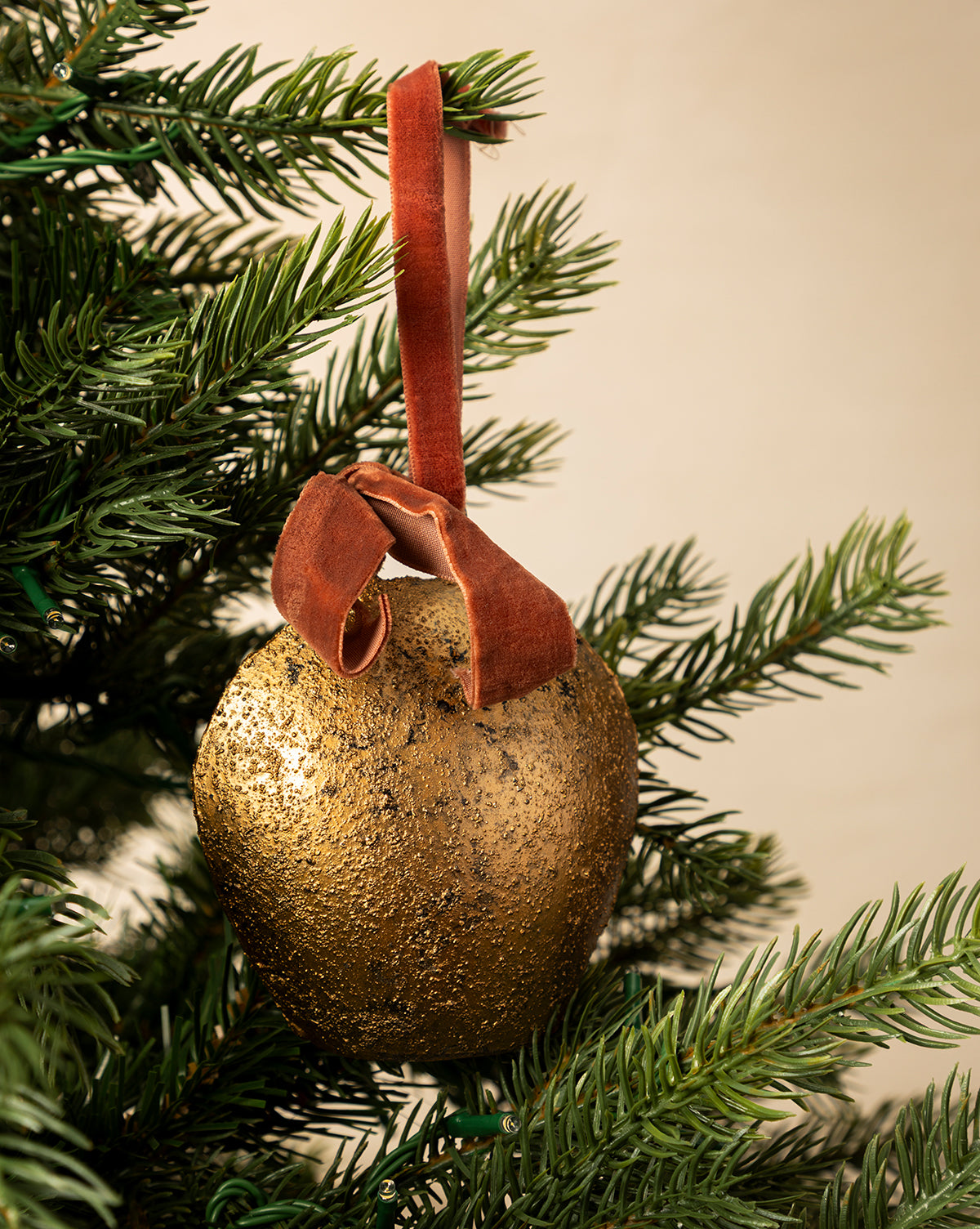The Antique Gold Metal Bell with Velvet Ribbon hangs on a green Christmas tree branch, adding elegant holiday charm to your décor with its antique finish and luxurious brown velvet ribbon, by McGee & Co.