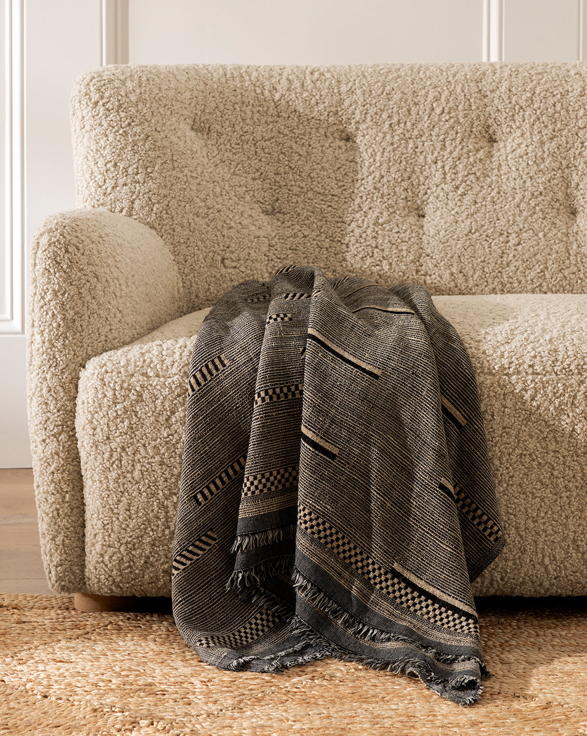 A textured beige sofa with the Anwar Woven Throw draped over one armrest, set on a woven rug in a bright, inviting room.