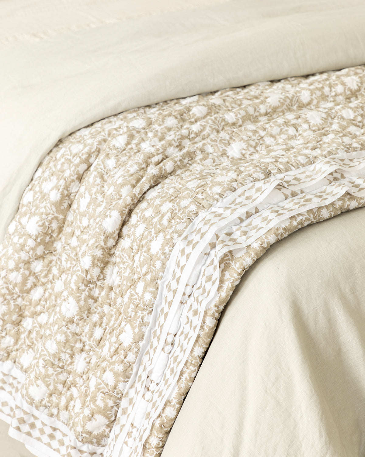 The Arcadia Block Print Kids Quilt, in beige and white with floral designs and a decorative border, is neatly folded at the foot of a light beige bed. Its soft, textured fabric adds comfort and style to any kids’ room.