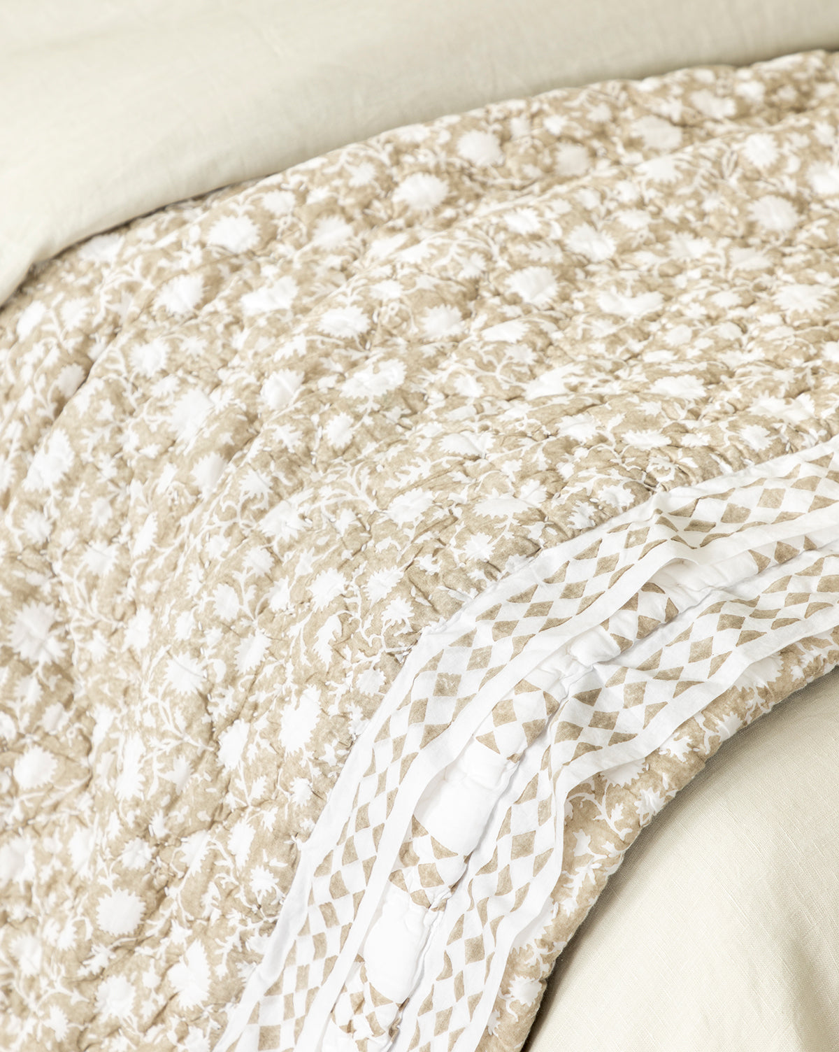 A close-up of the Arcadia Block Print Kids Quilt showcases its beige color, white floral design, and geometric border, layered over a light sheet on a bed.