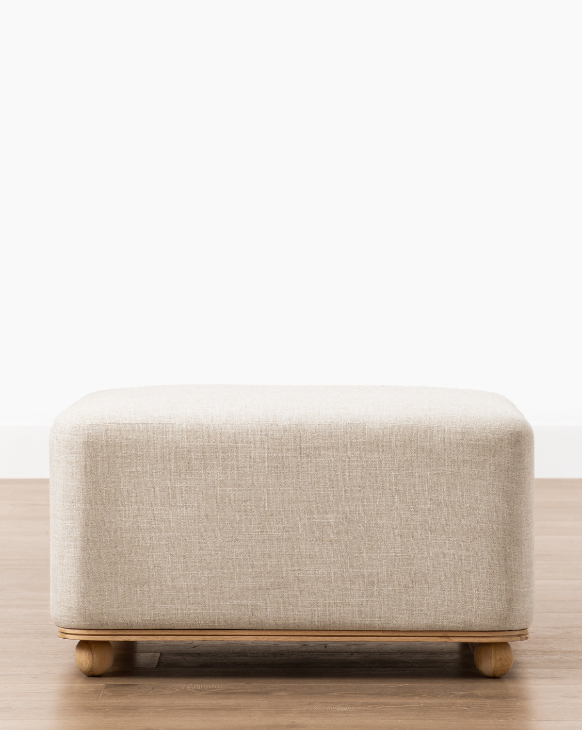 The Arie Ottoman, featuring a rectangular beige upholstered design with rounded corners and an oak frame, sits on a wooden floor against a white wall—an ideal choice for modern decor, by McGee & Co.